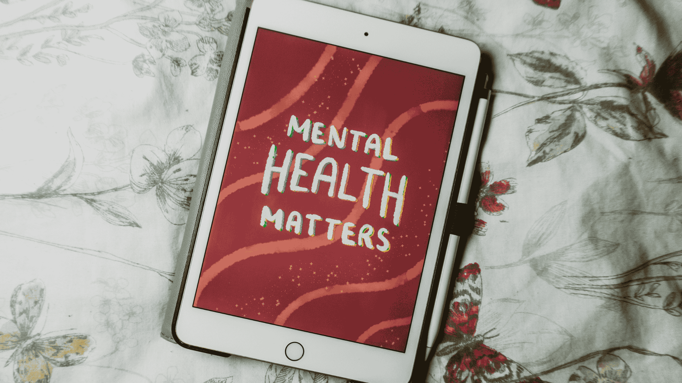 A tablet displaying a hand-lettered 'Mental Health Matters' illustration on a red background, resting on floral bedding representing the importance of journaling prompts for mental health