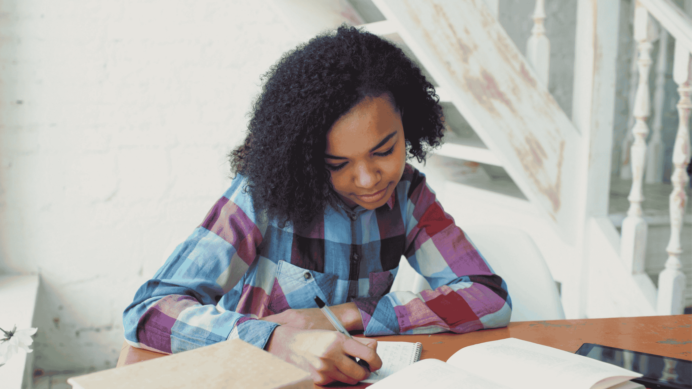 A teenage girl with curly hair writing in a journal at a bright wooden desk, with an open book and tablet nearby illustrating how journaling prompts for teens encourage self-reflection and academic growth