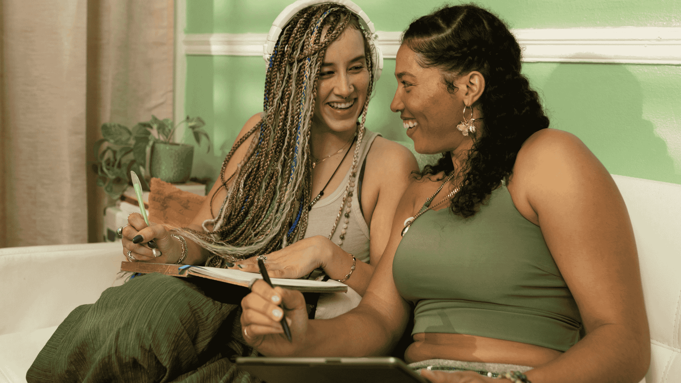 Two women laughing and writing in journals together on a couch in a green-toned room illustrating how sharing gratitude journal prompts can build connection and joy